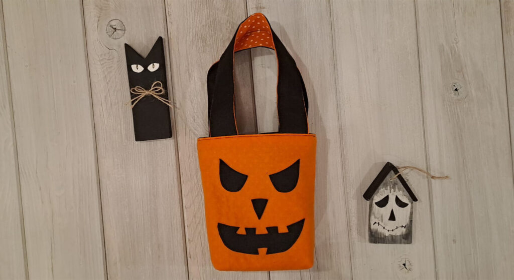 Borsetta ultimata, con la faccia di una zucca per festeggiare Halloween. A fianco un gattino nero e una casetta spettrale, in legno, realizzati a mano