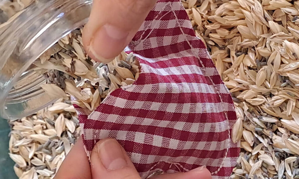 Dettaglio di mani che riempiono un cuoricino con lavanda e pula di farro con un vasetto di vetro.